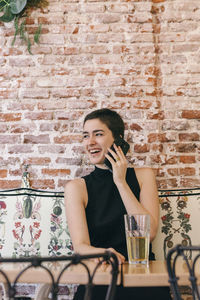 Young woman using phone while sitting on table against brick wall