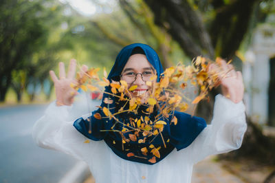 Portrait of happy young woman throwing autumn leaves on footpath