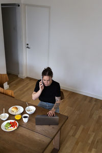 High angle view of young businesswoman talking on mobile phone while using laptop at dining table