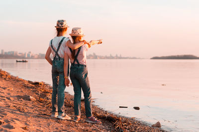 Two girls sisters stand on the banks of the river at sunset and look into the distance