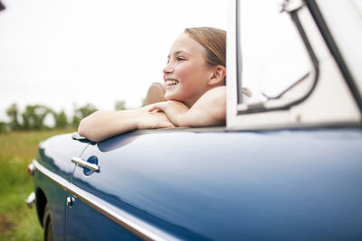 Happy girl sitting in car at field against sky