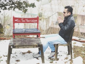 Side view of young man sitting on bench in snow