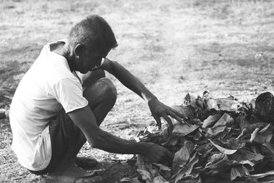 Side view of man crouching on field by heap of leaves