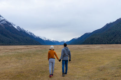 Rear view of couple holding hands walking on field against sky