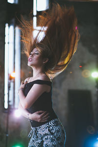 Side view of young woman with tousled hair standing against illuminated light