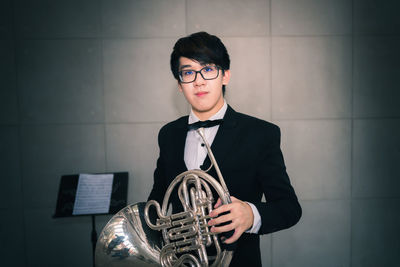 Portrait of man holding french horn while standing against wall