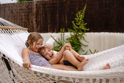 Cute barefooted little sister and brother hugging while lying in hammock and having fun during summer holidays