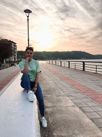 Full length portrait of young man sitting on footpath at sunset