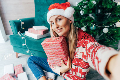 Portrait of cheerful woman wearing santa hat holding gift box while sitting by christmas tree at home