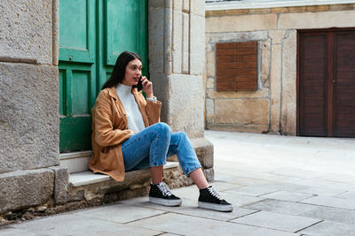 Full length of woman sitting on sidewalk against building