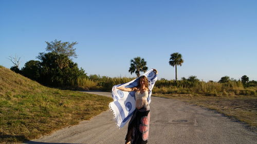 Young woman holding scarf standing on road against clear blue sky during sunny day