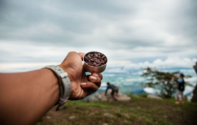 Cropped hand of woman holding roasted coffee beans against cloudy sky