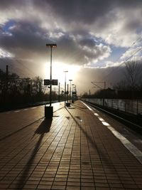 Empty footpath by street against sky during sunset