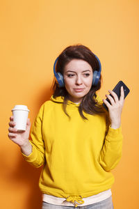 Young woman holding mobile phone while standing against yellow background