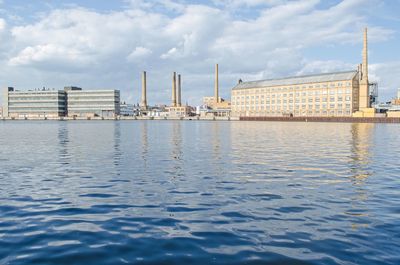 Industries in front of river against cloudy sky on sunny day