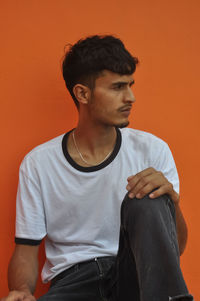 A handsome indian young guy looking away while sitting outside against orange wall with copy space