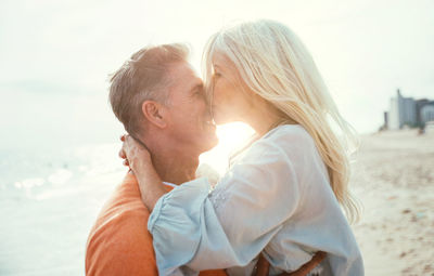 Side view of woman kissing man on nose at beach against sky