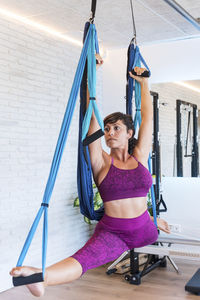 Slim woman doing splits with raised arms on aerial silks while practicing pilates against brick wall in gym