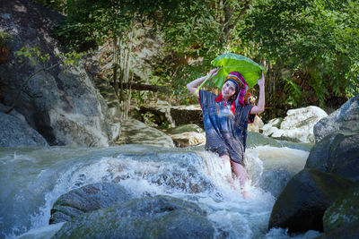 Portrait of smiling woman with leaf sitting on rock in river in forest