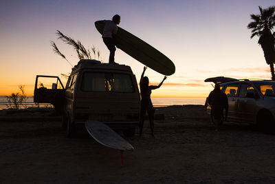 Silhouette man passing surfboard to woman while standing on mini van at san onofre state beach