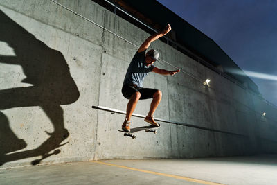 Low angle of talented male skater jumping with skateboard while doing stunts and training in illuminated skate park at night