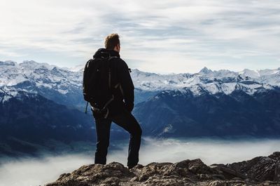 Rear view of man with backpack standing on rock against snowcapped mountains