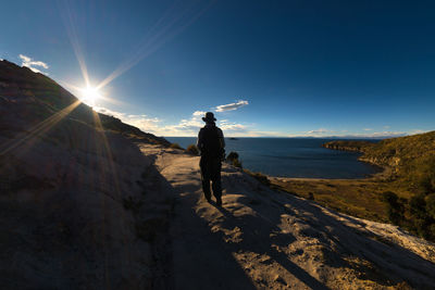 Rear view of hiker walking on cliff by sea against sky on sunny day