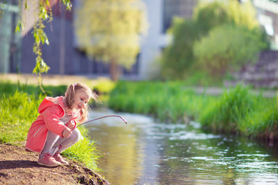 Side view of girl holding stick while crouching by stream