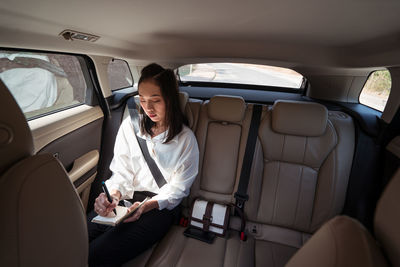 From above serious female in formal wear writing in notepad while riding on passenger backseat in comfortable cab