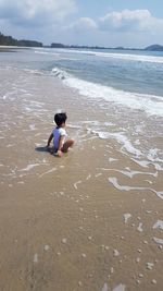 Side view of girl crouching at shore against sky during sunny day