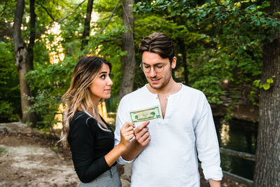 Young couple holding paper currency while standing against trees