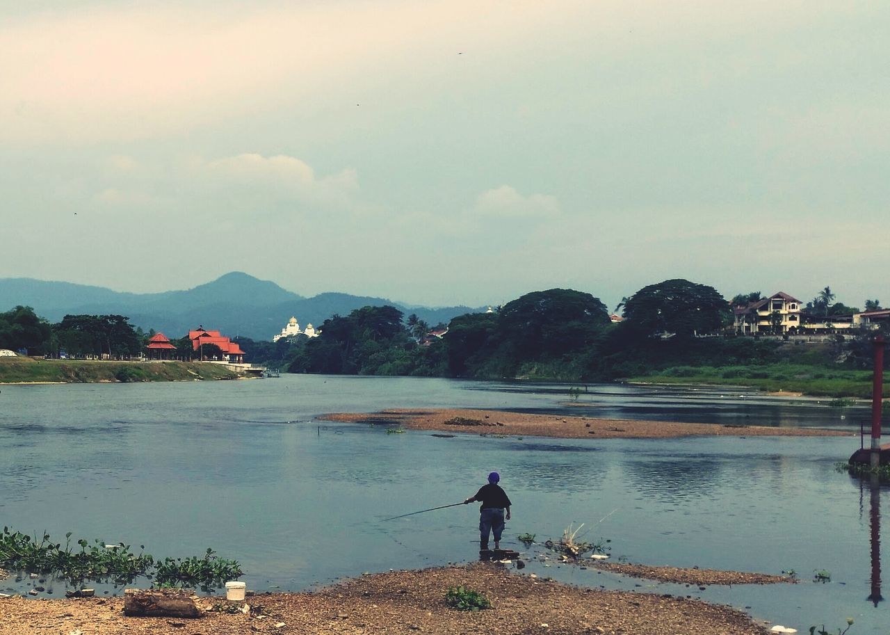MAN FISHING AT RIVERBANK AGAINST SKY