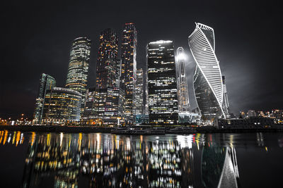 Illuminated modern buildings by river against sky at night