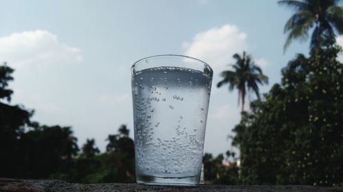 Close-up of glass of water with trees against sky