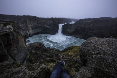 Low section of man sitting on cliff against waterfall