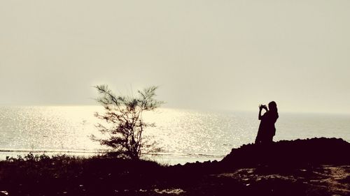 Silhouette woman photographing sea against clear sky