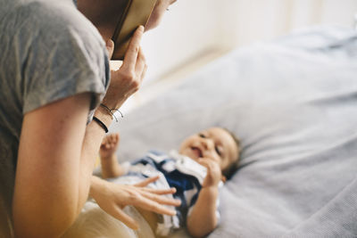 Cropped image of mother talking on mobile phone dressing son at bed