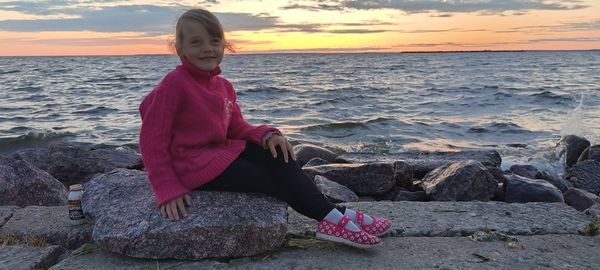 Woman sitting on rock by sea against sky during sunset