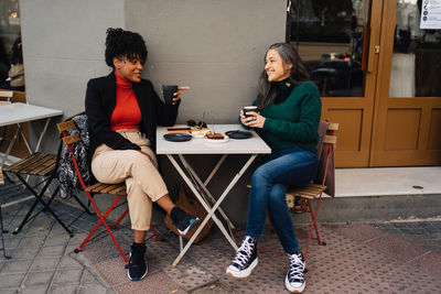 Smiling hispanic woman sitting at table with cakes and drinking hot latte during meeting with african american girlfriend in outdoor coffee shop