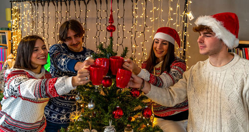 Group of four young friends toasting with red mugs in front of a christmas tree full of ornaments