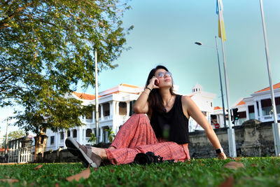 Smiling young woman sitting on grass against building and sky
