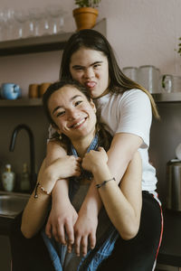 Young woman sticking out tongue with down syndrome hugging sister from behind in kitchen at home