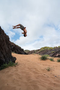 Side view of shirtless male leaping from cliff on sandy beach against cloudy blue sky on sunny summer day