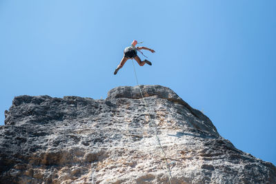 Low angle view of man rock climbing against clear blue sky