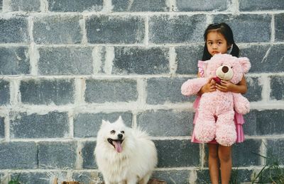 Portrait of cute girl with toy and dog standing against brick wall