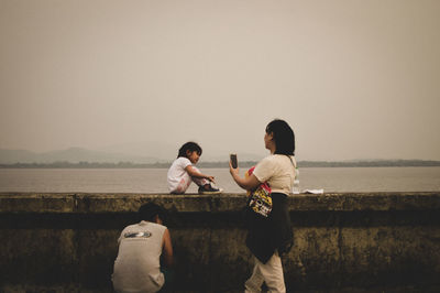 Rear view of men sitting on shore against clear sky