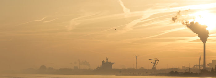 Smoke emitting from factory in city by river against sky during sunset