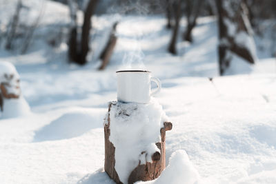 An enamelled mug with a hot drink standing on a snow-covered stump in a winter forest. steam rises