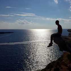 Side view of silhouette man sitting on cliff by sea against sky during sunset