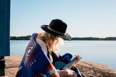 Woman sat on a rock playing the guitar on a sunny day by the water